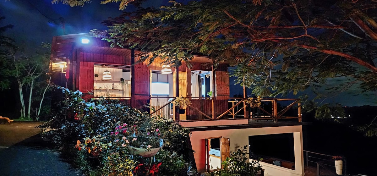 The rustic house is presented in low light, with wooden paneling and expansive windows. Surrounding greenery and colorful flowers enhance the natural ambiance. The structure features an elevated deck with a railing and invites a sense of connection to the outdoors.