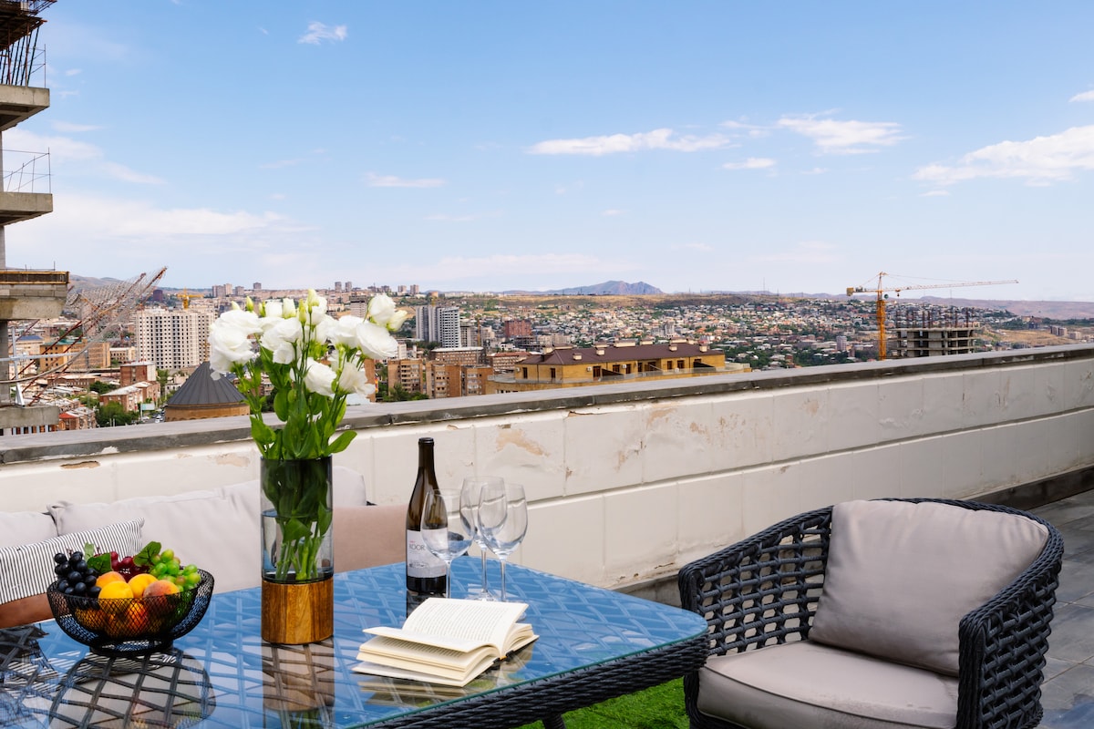 An outdoor terrace displays a modern glass table set with a bottle of wine, glasses, and an open book. A vase holds fresh white flowers, while a bowl of fruit adds color. The panoramic city view is visible in the background against a clear sky.