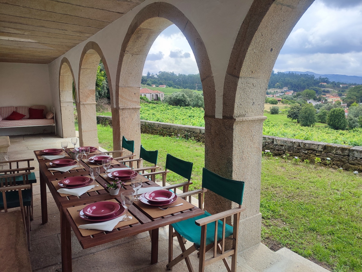 An outdoor dining area features a long wooden table set with plates and cutlery, surrounded by green chairs. Arched stone columns frame the space, providing views of the surrounding vineyards and hills, creating a setting for meals and relaxation in nature.