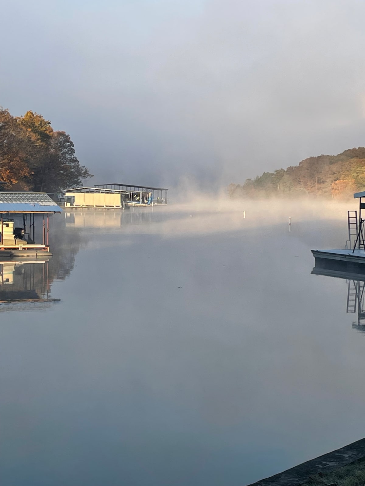 A tranquil lake scene is captured in the early morning, with calm waters reflecting the mist. Two docks with covered boats are visible, framed by gentle trees in autumn hues, creating a serene atmosphere along the quiet cove.
