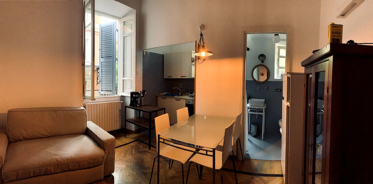 A cozy living area features a beige sofa and a glass dining table surrounded by four chairs. Natural light enters through the large window with blue shutters, illuminating the space. The kitchen and bathroom are accessible through an archway, presenting a streamlined layout.