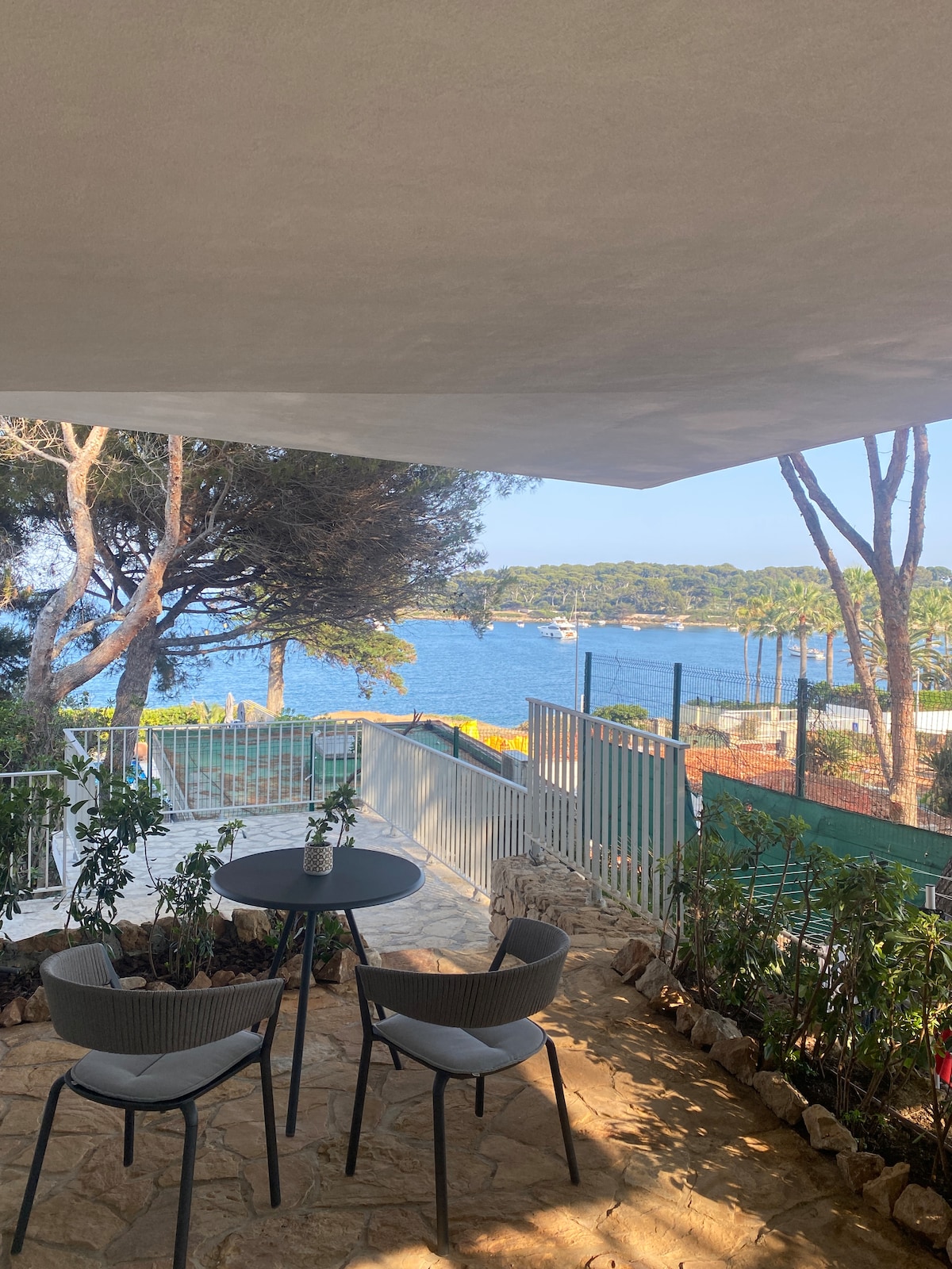 A private terrace features two chairs and a small table, surrounded by plants. The space overlooks the bay, with views of boats in the water and lush greenery in the background.
