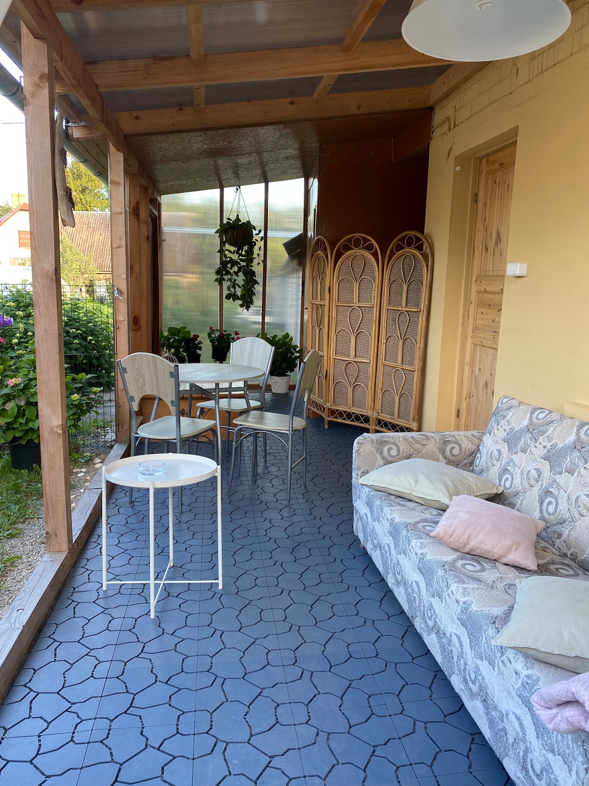A sheltered seating area with a patterned floor features a light-colored couch with soft pillows and a small round table surrounded by four chairs. A folding screen stands nearby, and sunlight filters through the open space, illuminating the greenery outside.