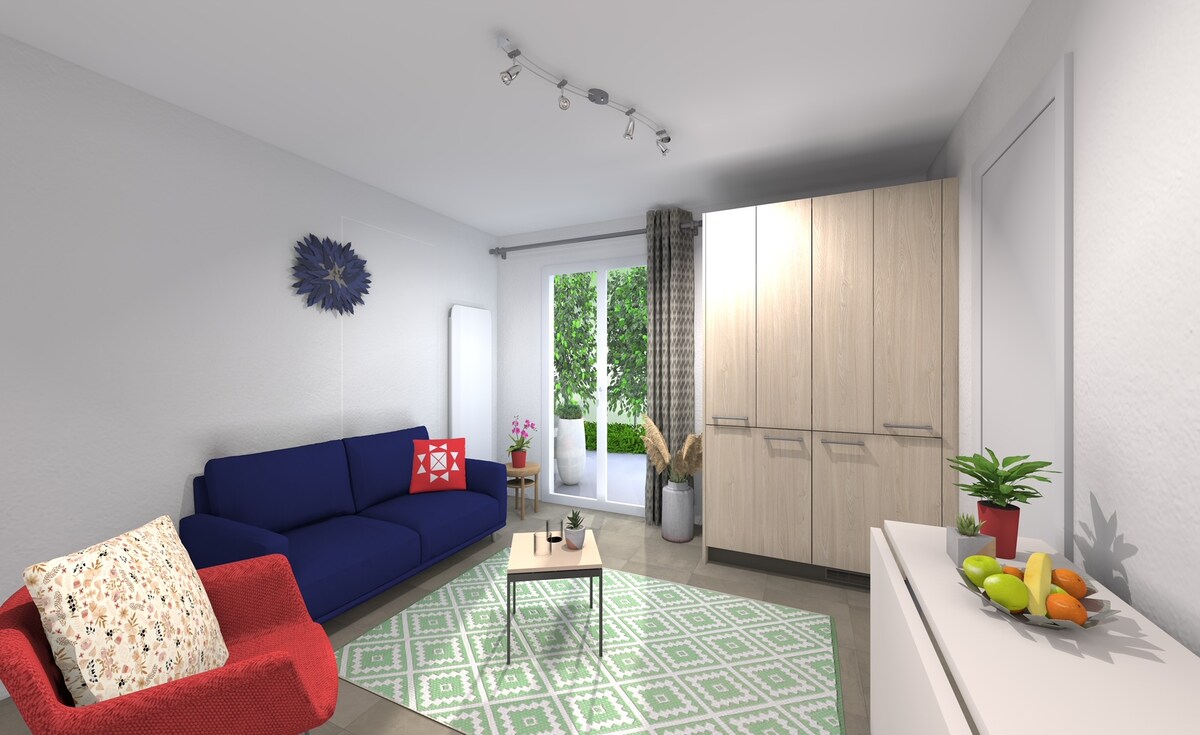 The living room features a blue sofa and a red armchair, accompanied by a small coffee table arranged on a patterned rug. Natural light enters through glass doors leading to a green space, while a plant and decorative items add a touch of warmth to the room.
