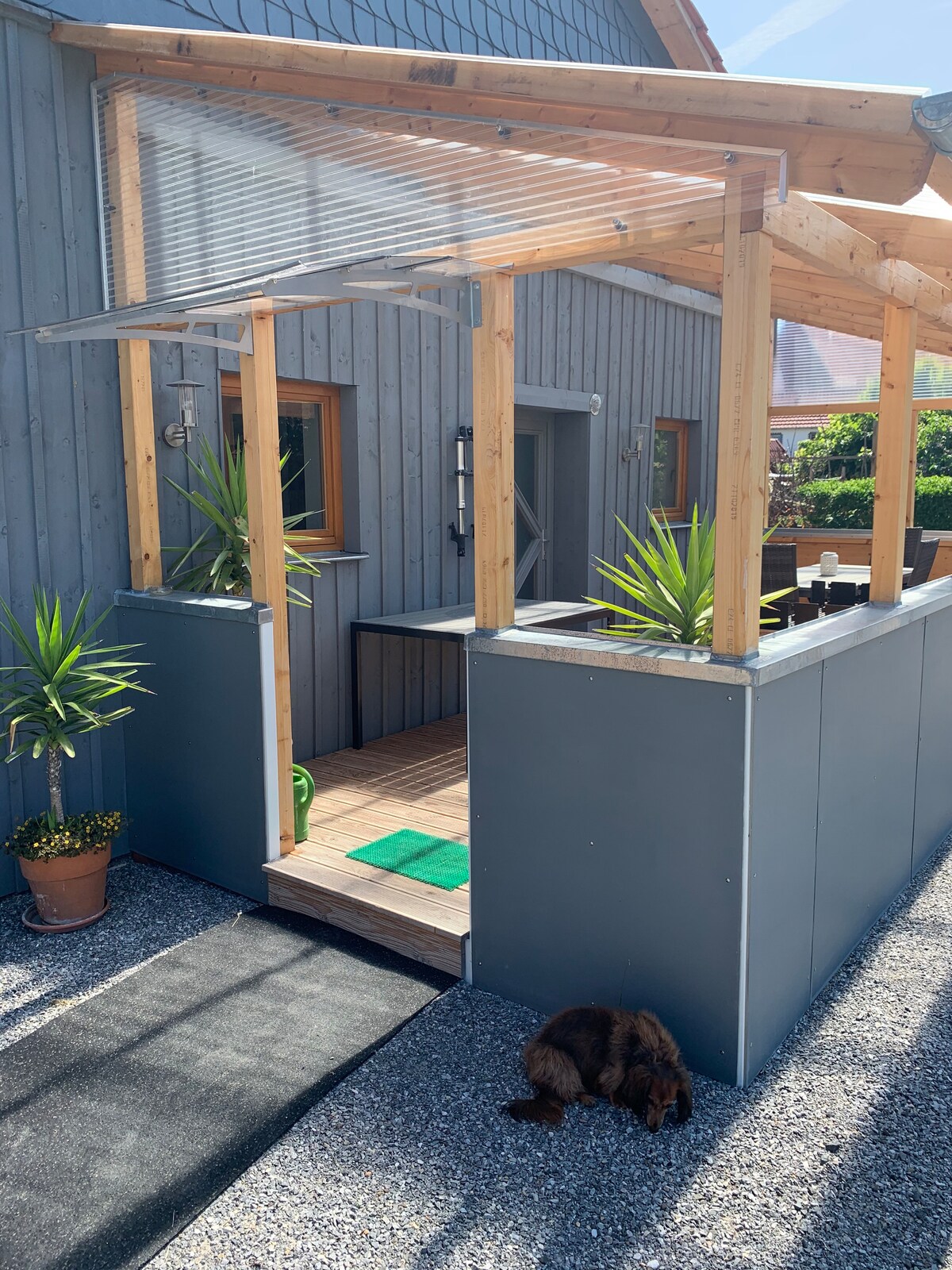 An entrance pathway is presented, bordered by modern planters with greenery. The wooden overhang provides shelter over a deck area, which features light-colored flooring. A small animal rests quietly on the gravel, contributing to the tranquil ambiance of the outdoor space.