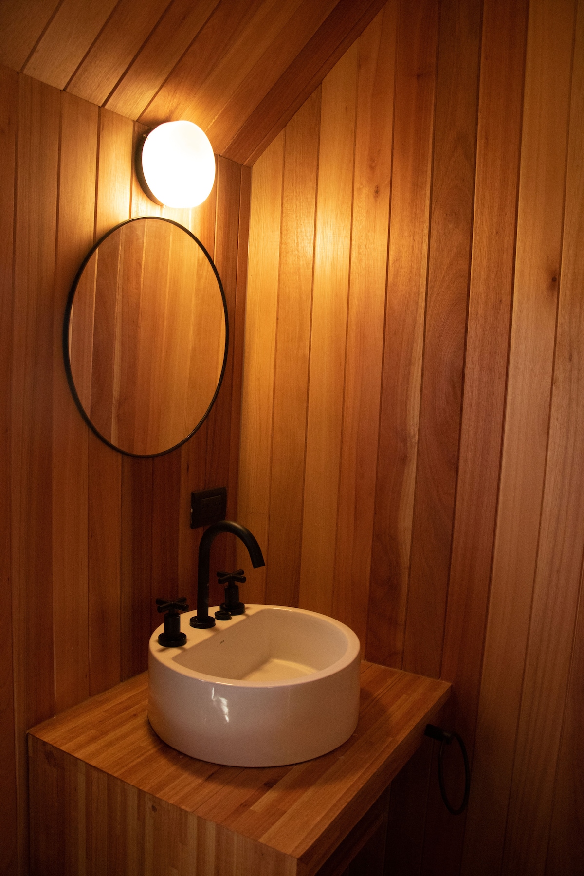 A modern bathroom features warm wooden walls creating a natural ambiance. A sleek white sink is paired with matte black fixtures and is placed on a wooden countertop. A round mirror reflects soft light from an overhead fixture, enhancing the minimalist design.