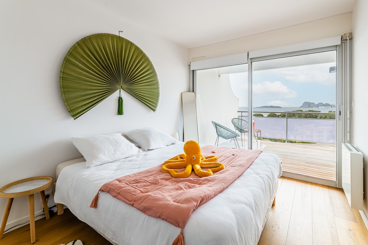A spacious bedroom features a large bed covered with a light blanket, accented by an orange plush octopus. Natural light floods in through sliding glass doors that open to a terrace with scenic views. A decorative fan hangs above the bed, adding a unique touch.