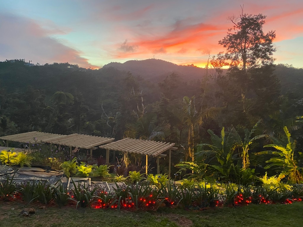 A sunset view over the nearby mountains is captured, with vibrant colors illuminating the sky. A pergola structure is visible in the foreground, surrounded by lush greenery. Soft lights are placed along the ground, enhancing the natural setting.