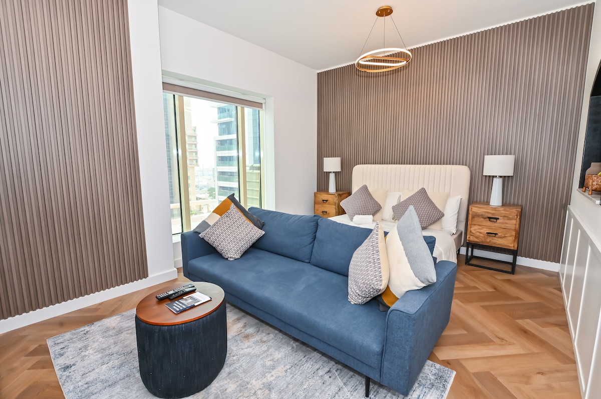 A modern living area is displayed with a plush blue sofa adorned with assorted decorative pillows. A circular table rests nearby, positioned on a soft area rug. A spacious bed is visible in the background, complemented by bedside tables and warm lighting from stylish lamps.