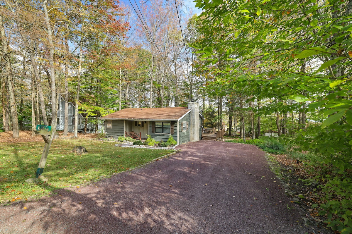 The exterior view of the house showcases a landscaped front yard with greenery and a gravel driveway leading up to the entrance. Surrounding trees in varied autumn colors create a natural, serene setting.