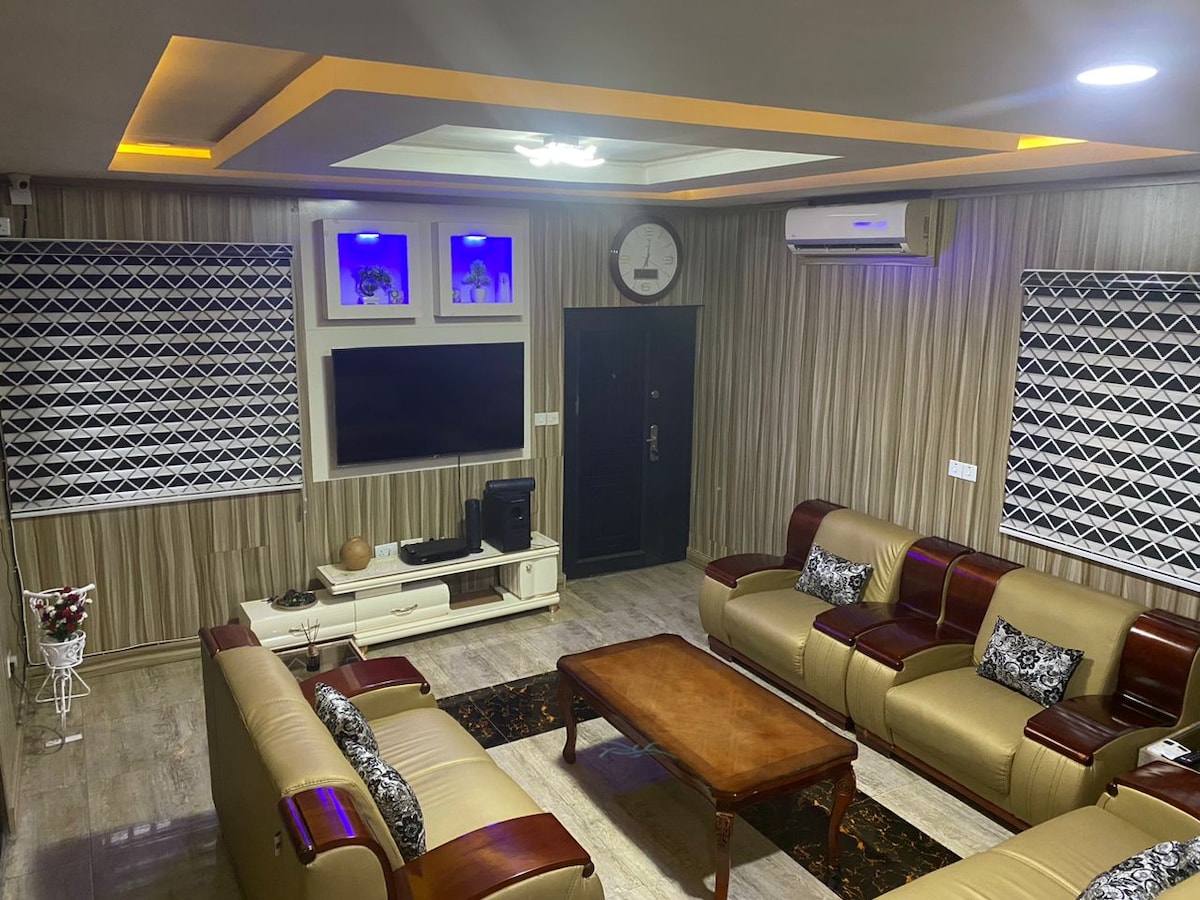 A spacious living area is featured, equipped with comfortable seating and a wooden coffee table at the center. Wall-mounted artwork is displayed, and a television is positioned opposite the seating. Soft lighting enhances the modern décor, while air conditioning units are visible high on the walls.