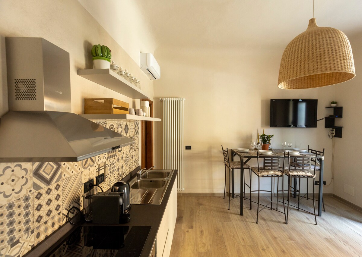 A modern kitchen is equipped with stainless steel appliances, including a stovetop and a kettle. A dining table with metal chairs is visible in the background, accompanied by a wall-mounted television. Natural light enters through a window, enhancing the light wood flooring.