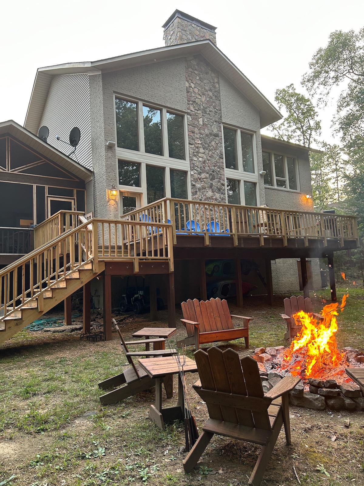 The exterior of the house features a stone accent wall and large windows, allowing ample natural light. A deck with outdoor seating overlooks a fire pit surrounded by wooden chairs. Lush greenery and a gravel path enhance the natural surroundings.