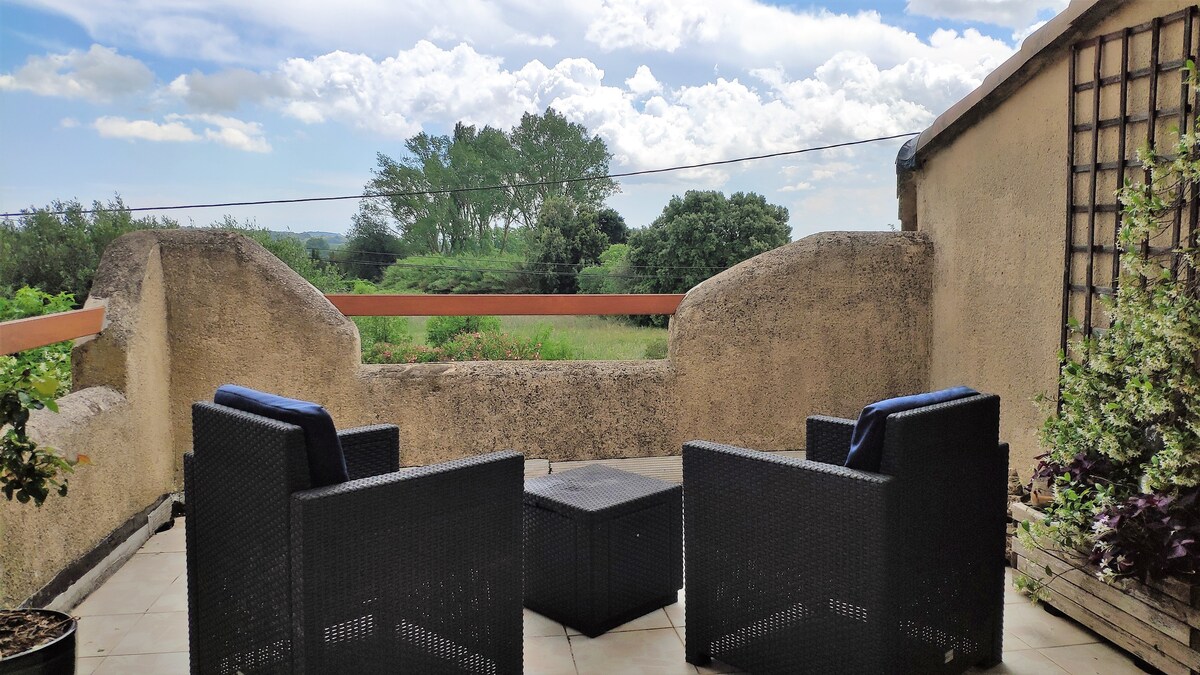 A terrace is presented with stylish outdoor seating, including two chairs and a small table. The view features lush greenery and open skies, providing a serene backdrop. The walls are textured, contributing to the cozy atmosphere of the space.