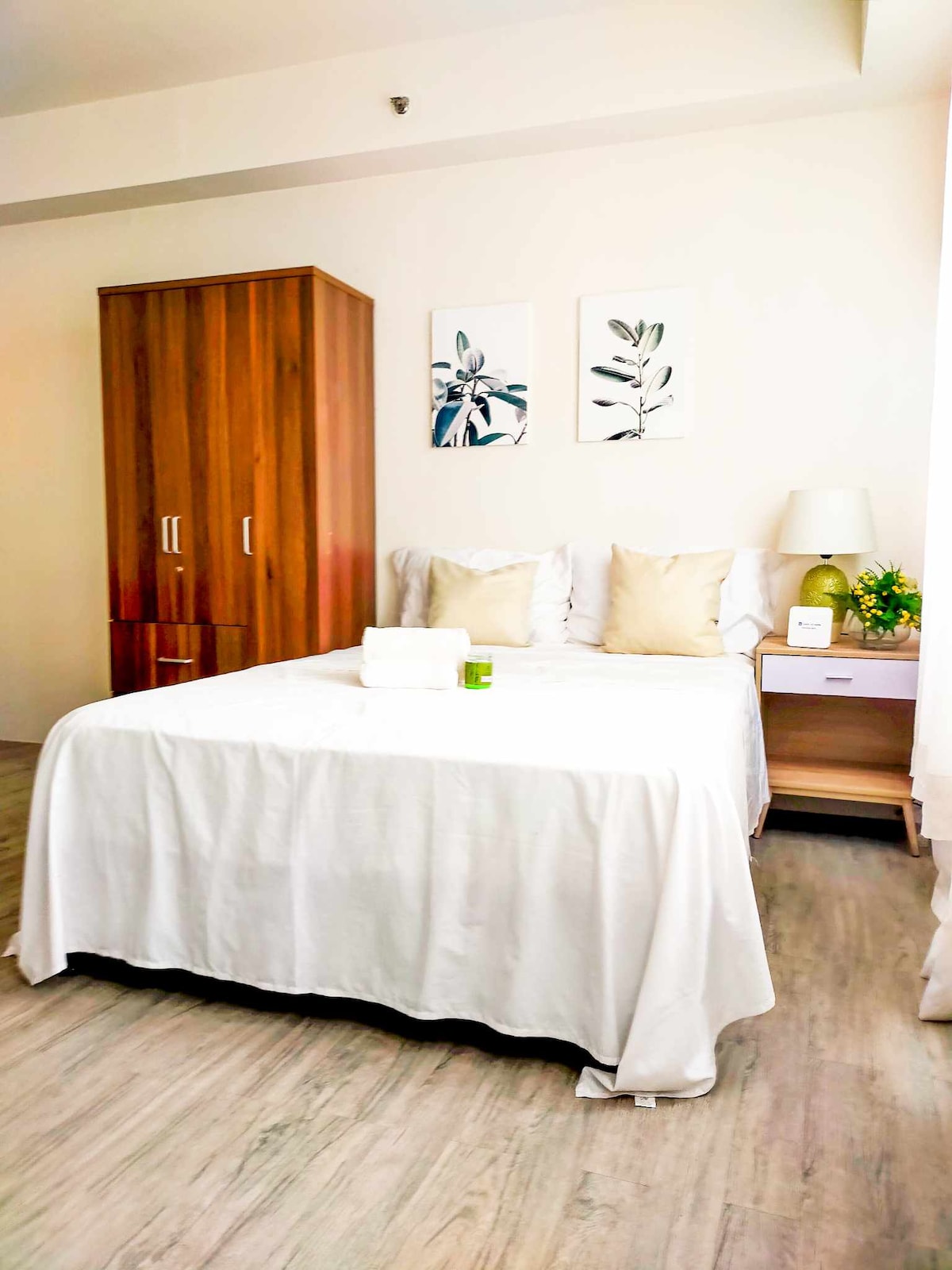 A spacious sleeping area is presented with a large bed adorned with a white coverlet. Two decorative pillows are placed at the head of the bed. A wooden wardrobe and nightstand with a lamp are visible, accompanied by framed botanical artwork on the wall.