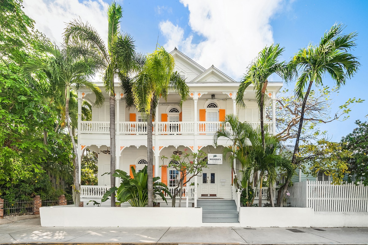 The historic Victorian building features a two-story facade with vibrant orange shutters and white trim. Lush palm trees and greenery line the entrance, with a white picket fence visible in the background. A welcoming porch showcases intricate railings and multiple entrances.