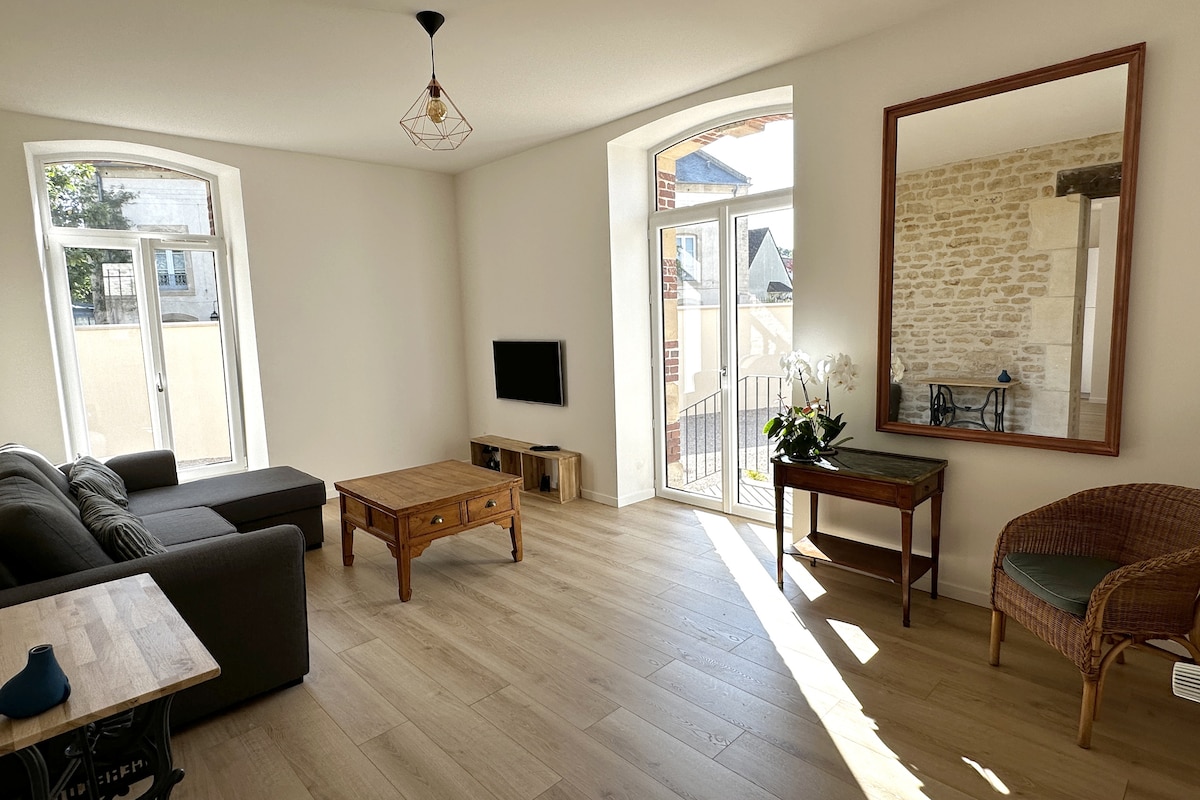 The living area is bright and spacious, featuring large windows that allow natural light to fill the room. A comfortable sofa and wooden coffee table are positioned on a light wood floor. A decorative mirror and a wicker chair add to the inviting decor.