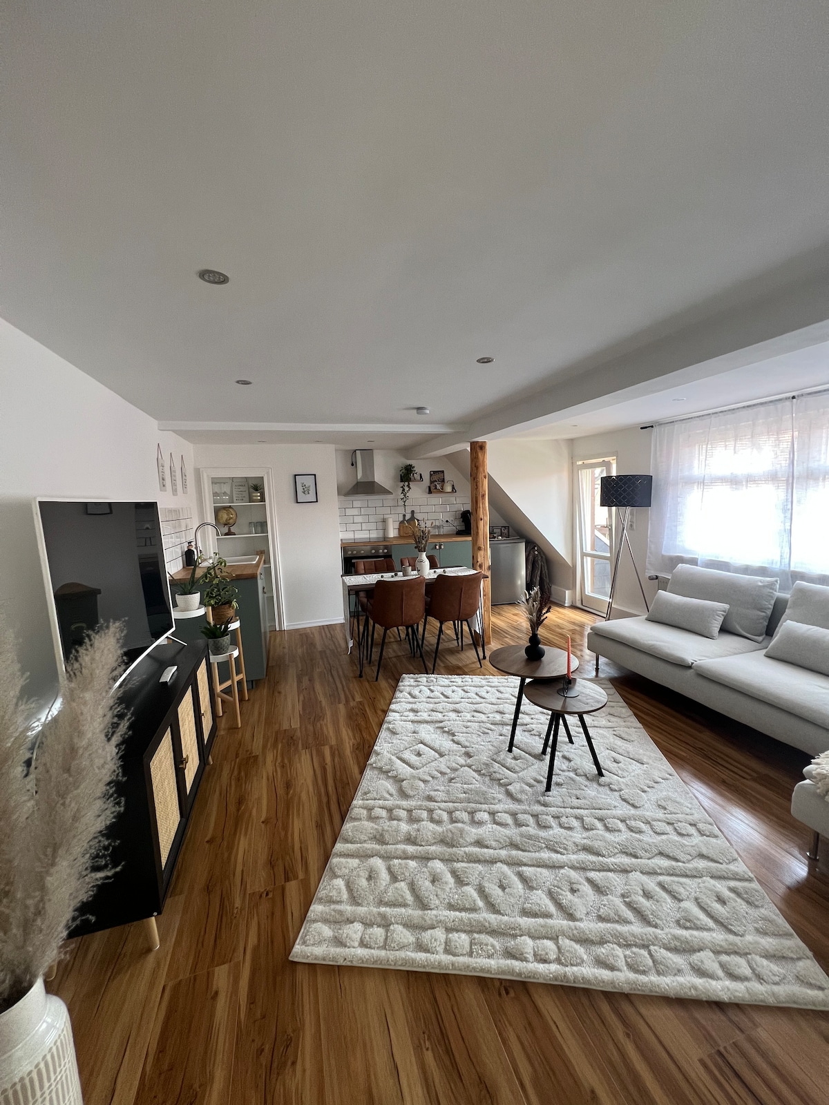 An inviting living space showcases a light, open layout. A comfortable sofa is positioned next to a small coffee table on a textured area rug. A dining area with four chairs is visible, accompanied by natural light streaming through large windows.