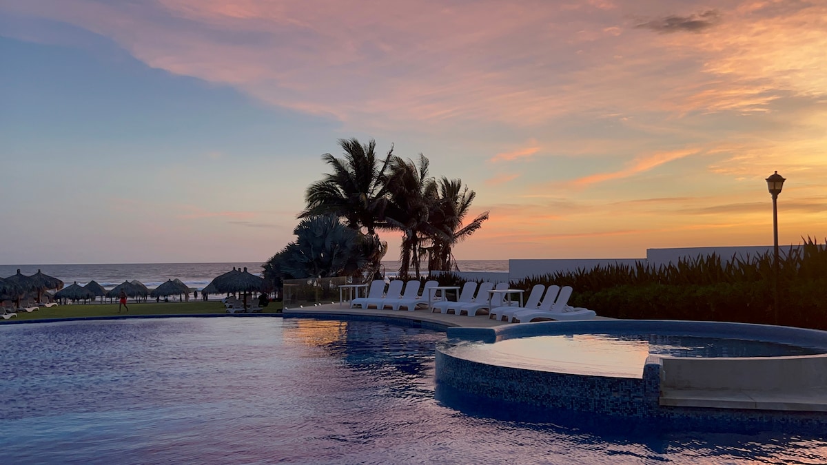 An inviting pool area is depicted, featuring a large swimming pool with a circular section. Sun loungers are positioned nearby, and palm trees gently sway against a colorful sunset sky. The ocean is visible in the background, creating a serene coastal atmosphere.