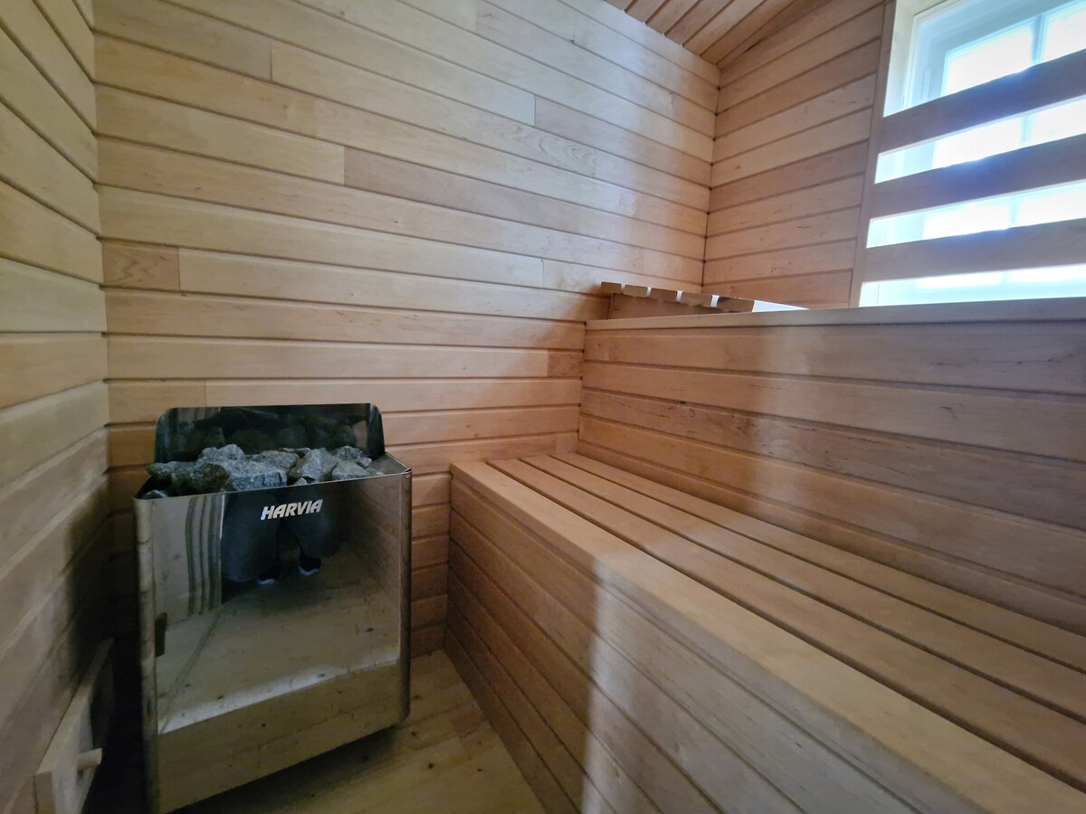 A wooden sauna room features a stone heating unit and spacious benches arranged against the walls. Natural light enters through a window, highlighting the warm tones of the wooden planks that create a relaxing atmosphere.