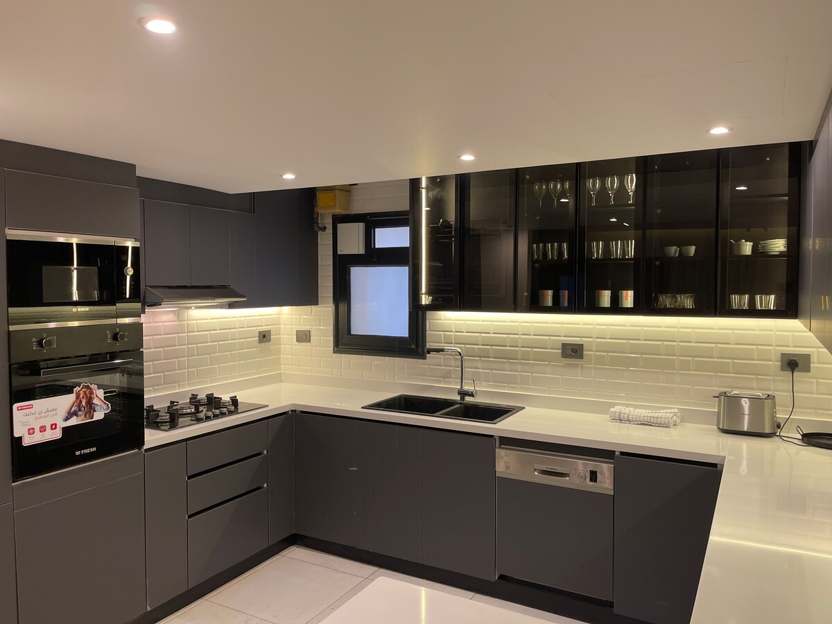 A modern kitchen is equipped with sleek black cabinetry and a minimalist design. Stainless steel appliances include an oven and a cooktop, while a glass-fronted cabinet displays various glassware. A double sink and ample counter space provide functionality and convenience.