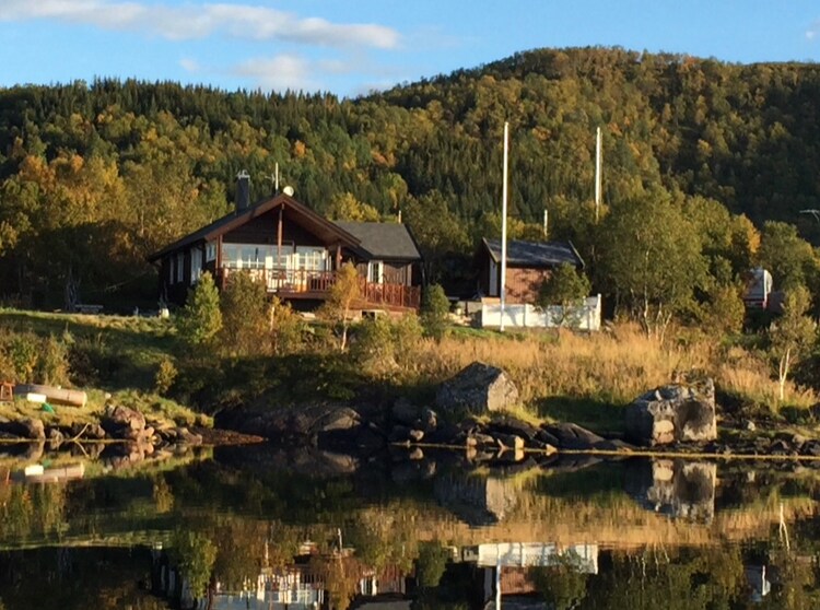 A unique cabin features large windows and a spacious outdoor deck, reflecting on calm waters. Surrounded by lush green hills and trees, this scenic location provides a serene atmosphere. Nearby structures complement the natural beauty, and the landscape is illuminated by soft sunlight.