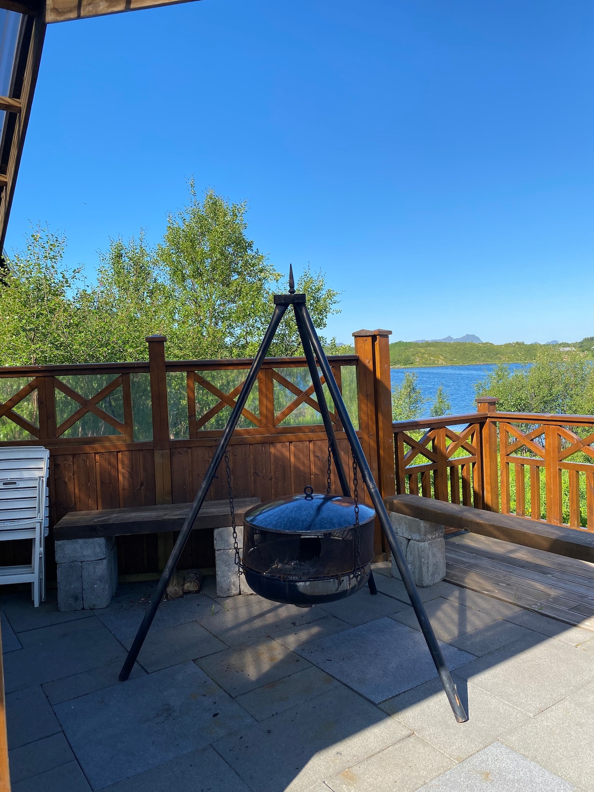 An outdoor seating area is framed by wooden railings, offering views of a serene waterway. A black cooking tripod stands prominently on the stone patio, surrounded by natural greenery. Clear blue skies enhance the inviting ambiance of the space.