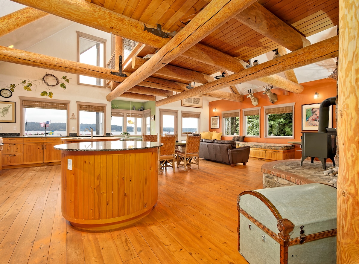 An open concept living area showcases wooden beams and warm-toned flooring. A circular kitchen island is surrounded by wicker chairs, while a cozy seating area features a couch and a wood-burning stove. Large windows provide views of the surrounding landscape.