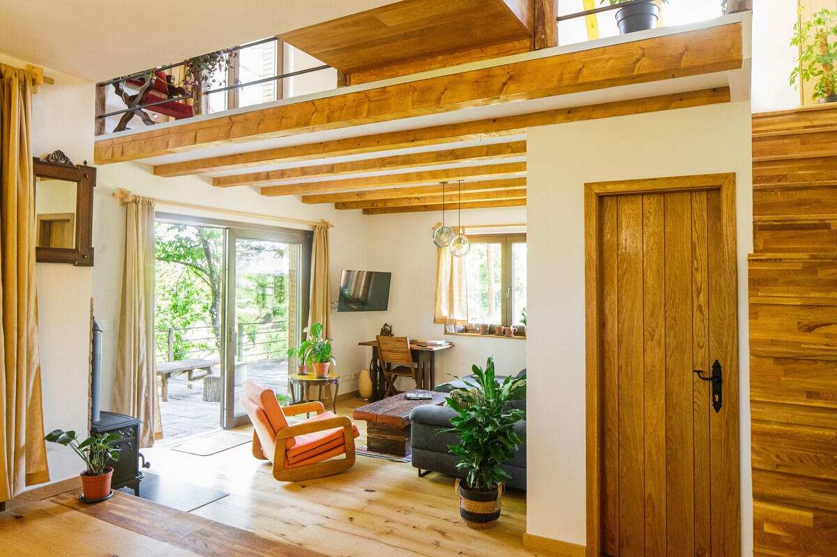 A welcoming living space features wooden beams and warm-toned flooring. Natural light floods in through large glass doors, revealing a view of the outdoors. A comfortable armchair and stylish sofa are positioned near a small table. Potted plants add greenery, enhancing the tranquil environment.
