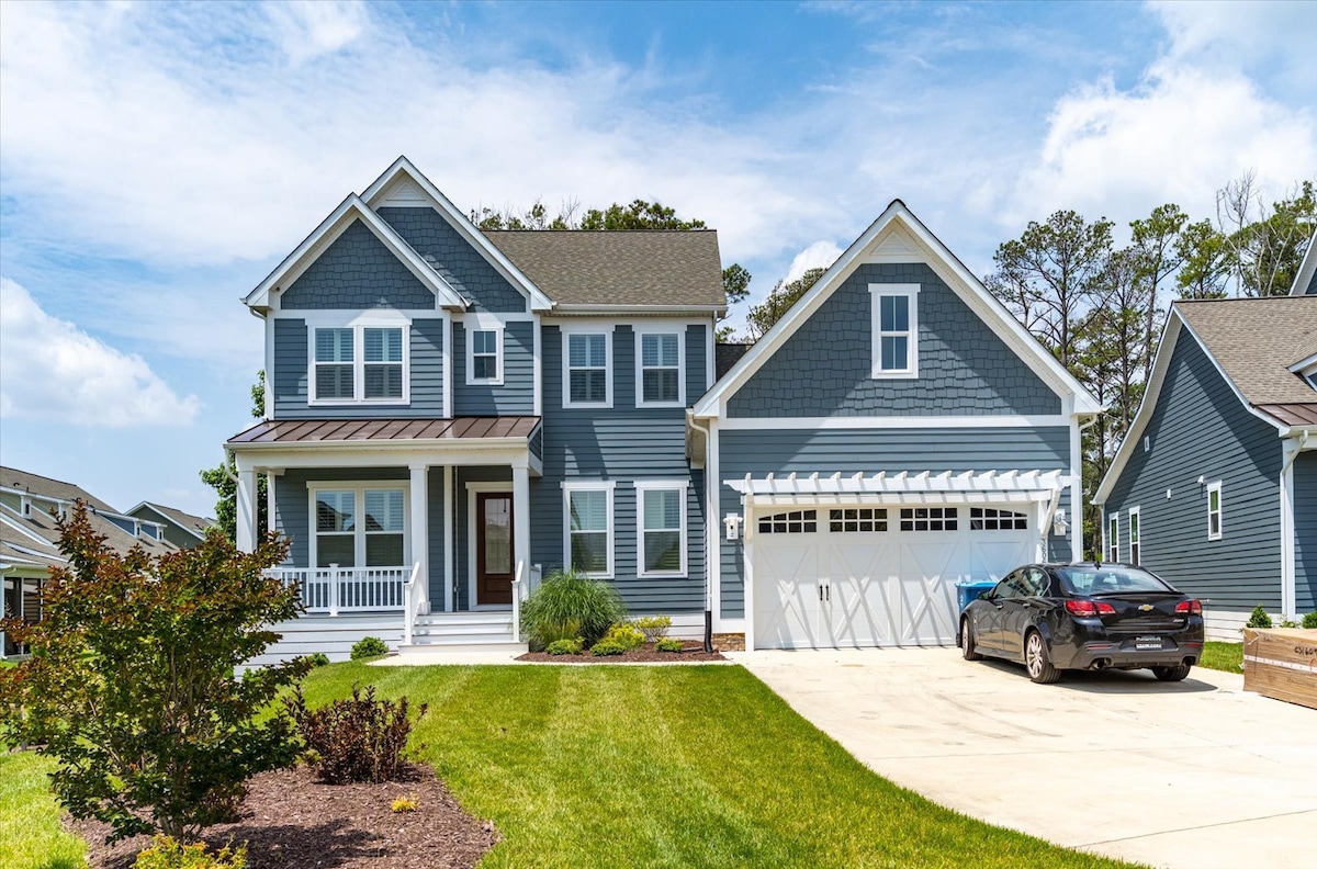 A two-story house is displayed with a modern design, featuring blue siding and white trim. A covered porch provides access to the front entrance, while a spacious driveway is visible, accommodating a parked car. Lush green lawn and shrubs surround the property.