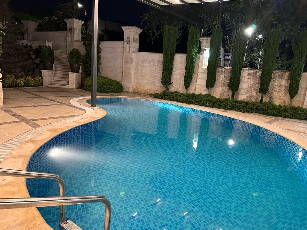 A serene outdoor pool area is featured, with a clear blue water surface reflecting surrounding lights. Smooth, tiled edges provide access to the pool, while neatly trimmed greenery and stone walls create a tranquil atmosphere, perfect for relaxation.