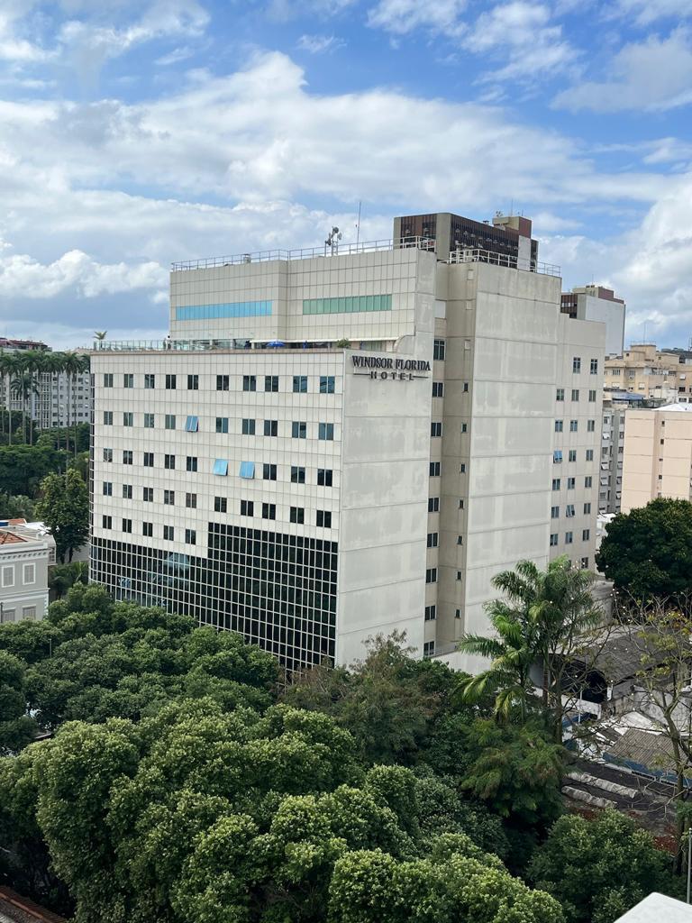 A multi-story building is viewed from above, showcasing a modern facade with large windows. Lush green trees surround the base, contrasting with the grey tones of the structure. A bright sky with scattered clouds can be seen in the background.