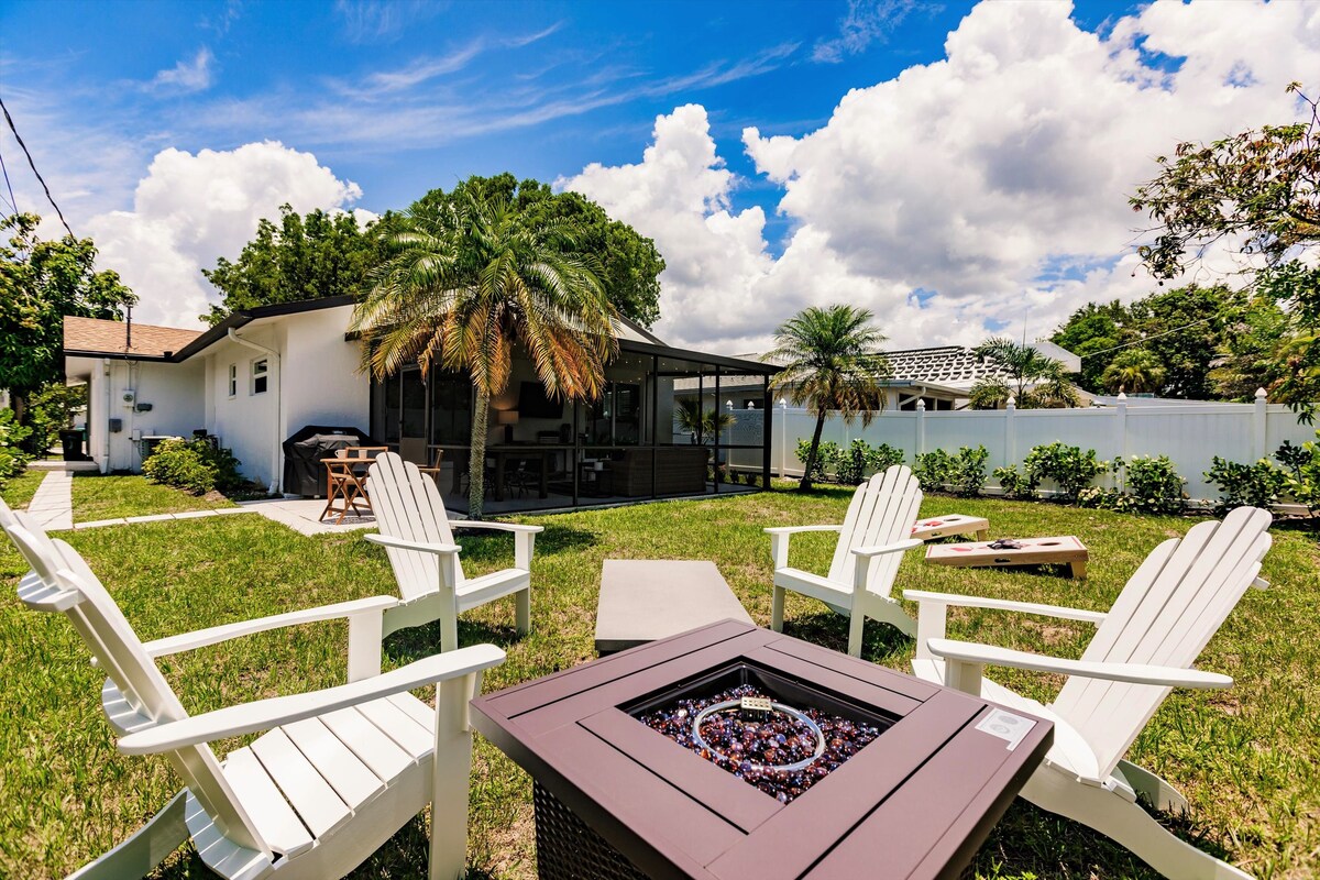An outdoor space is highlighted by a fire pit surrounded by four white adirondack chairs. The lush green yard features a path leading to a screened lanai. Palm trees and cloudy blue skies create a relaxing ambiance.