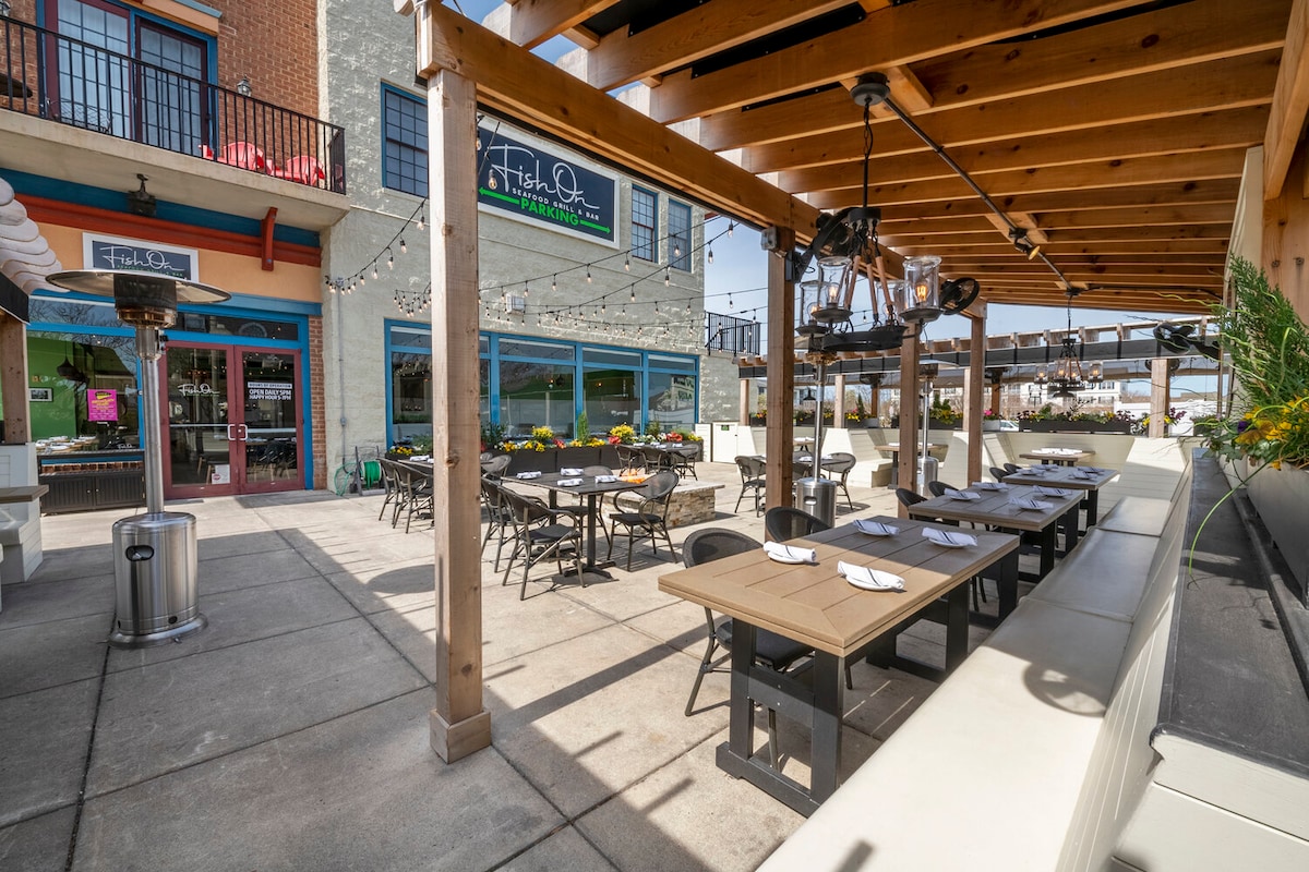 An outdoor dining area features a wooden pergola overhead, providing shade. Multiple tables are arranged with black chairs, and silver heating lamps are positioned above. Potted plants and flowers add color to the space, while neighboring storefronts are visible in the background.