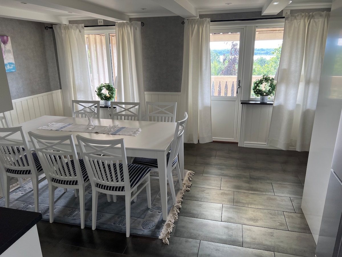 A dining area is featured with a long white table surrounded by eight chairs, positioned on a patterned rug. Large windows draped with light curtains allow natural light to illuminate the space, while greenery is placed on the table and windowsill, enhancing the inviting atmosphere.