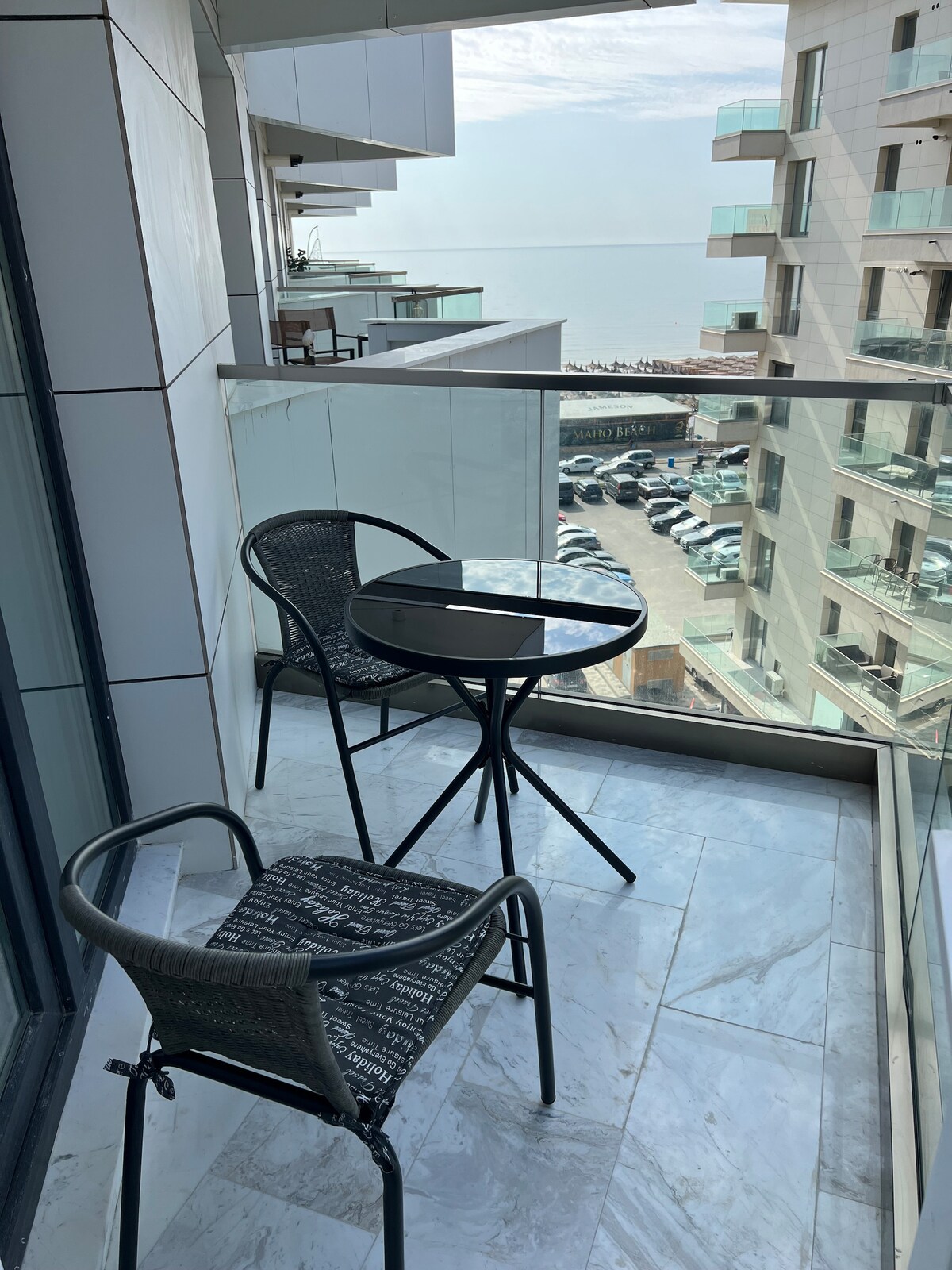 A balcony features a round table surrounded by two black chairs. The glass railing allows for an unobstructed view of the surroundings, including distant buildings and a glimpse of the water. The floor is covered in light-colored tiles, enhancing the outdoor space.