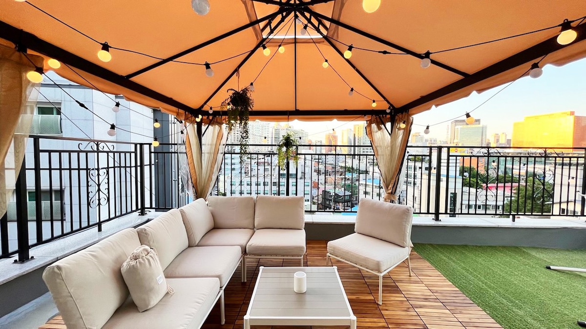 A spacious terrace is covered with a stylish canopy, featuring comfortable seating arrangements and a low table. Soft lighting hangs above, creating a cozy setting. The view captures a city skyline, providing a vibrant backdrop for relaxation.