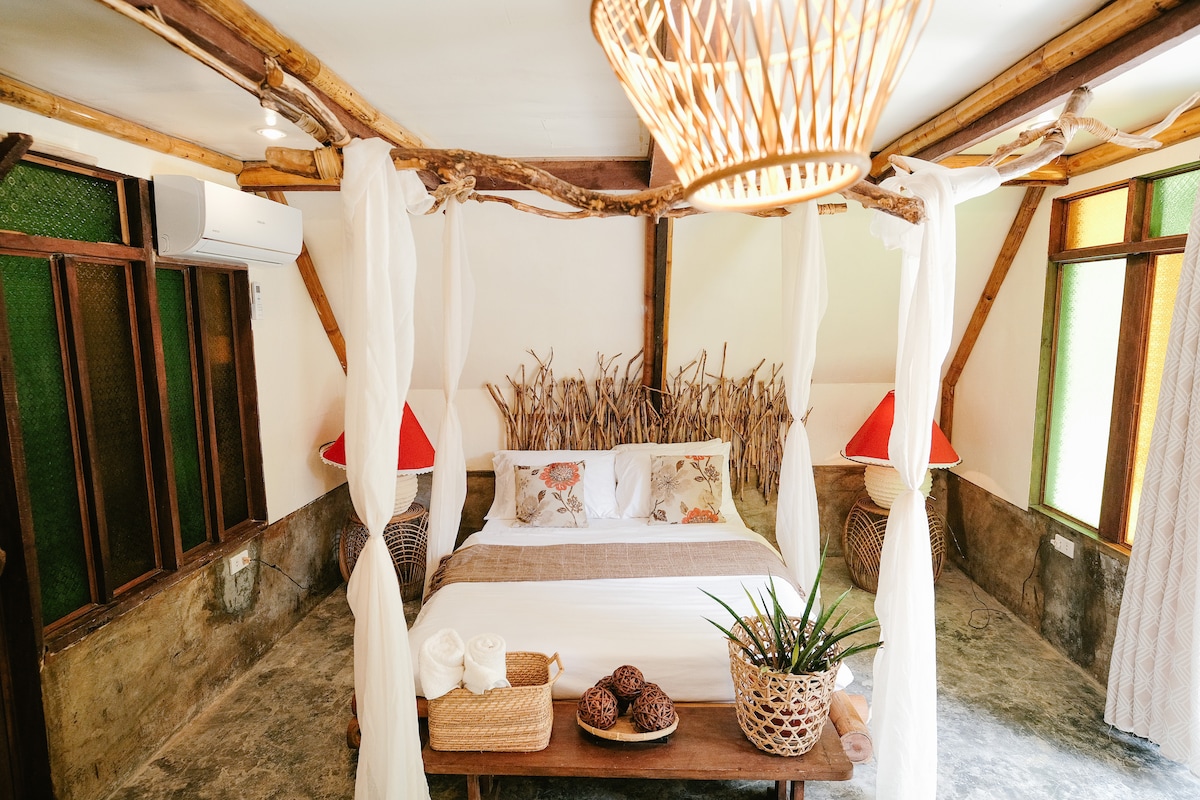 A spacious bedroom features a large bed surrounded by bamboo accents, complemented by light-colored bedding. Soft drapes create a cozy atmosphere. Two bedside lamps provide warm lighting, while a natural fiber rug adds texture. A wooden table holds decorative elements and fresh towels.