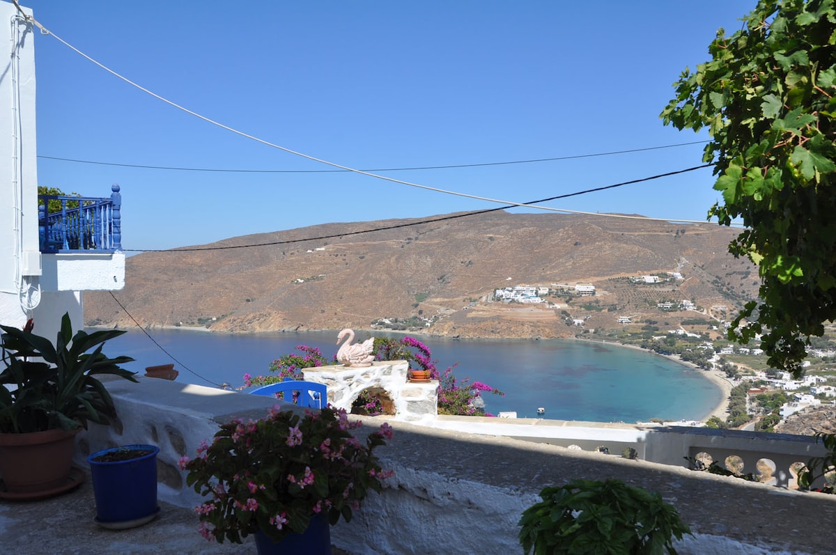 A panoramic view captures the serene coastline and hills of the sea, framed by blooming flowers and greenery. A small seating area, adorned with potted plants, invites relaxation. The sky is clear blue, enhancing the natural beauty of the landscape.