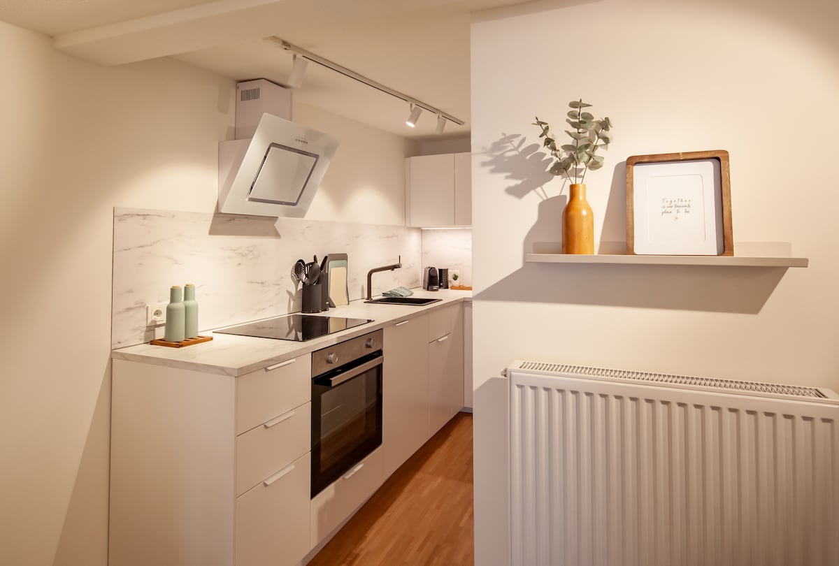 The kitchen features modern cabinetry with a smooth countertop, equipped with essential appliances. A minimalist shelf holds a decorative vase and framed artwork. Soft lighting highlights the warm wood flooring, creating a functional space for cooking and dining.