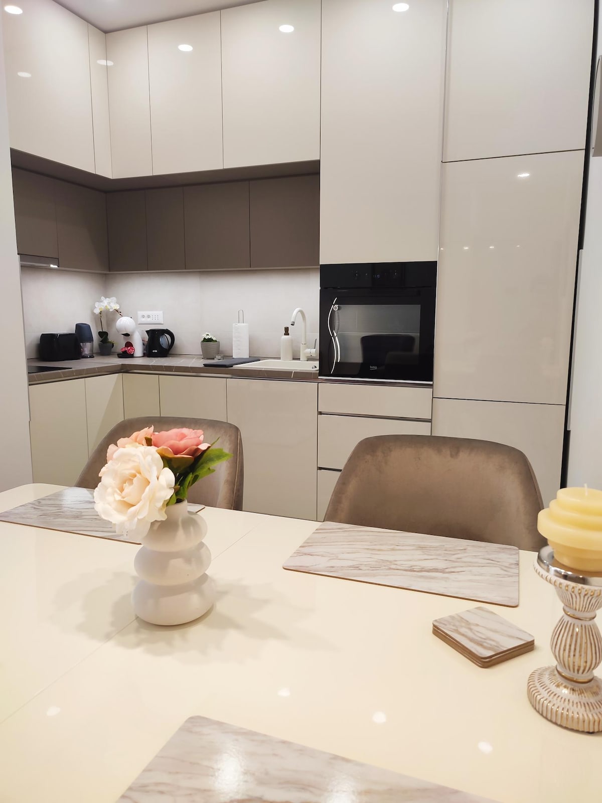 A modern kitchen is presented with sleek cabinetry and fitted appliances. A dining table is set with decorative placemats, featuring a simple vase holding fresh flowers. Soft lighting highlights the smooth surfaces and neutral color palette, creating a clean and contemporary look.