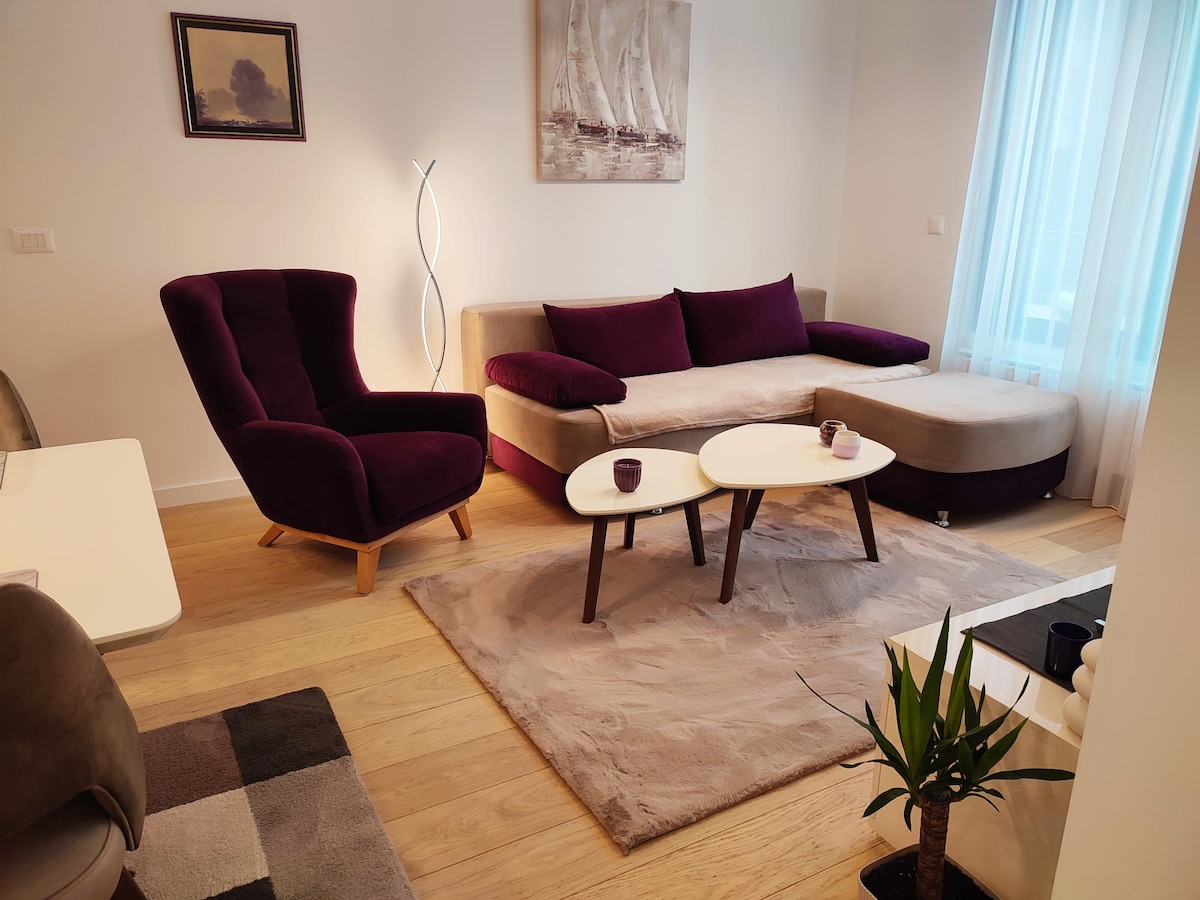 A modern living area is presented with a plush purple armchair and a curved sectional sofa in neutral tones. Two coffee tables are positioned on a soft area rug, while a potted plant adds a touch of greenery. Natural light filters in through sheer curtains.