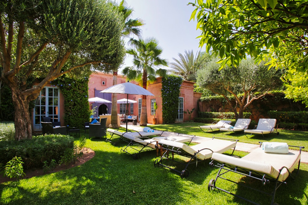 A spacious garden is displayed, featuring several sun loungers arranged on lush green grass. Large trees provide shade, while a patio area with outdoor seating is visible in the background, surrounded by well-maintained shrubbery. A large umbrella offers additional shade.