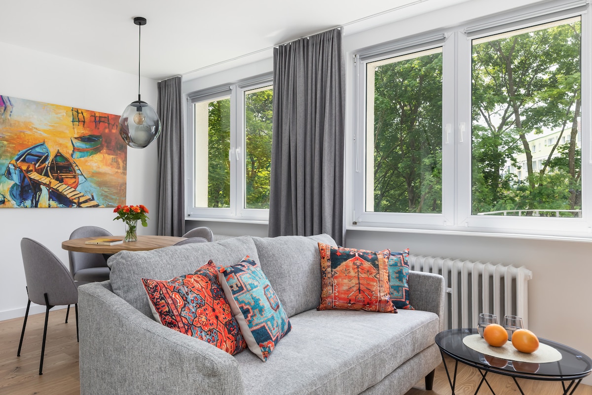 A light-filled living space features large windows framing views of lush greenery. A grey sofa adorned with colorful cushions sits alongside a glass table and modern dining chairs. A vibrant wall painting depicting boats adds a pop of color to the room.