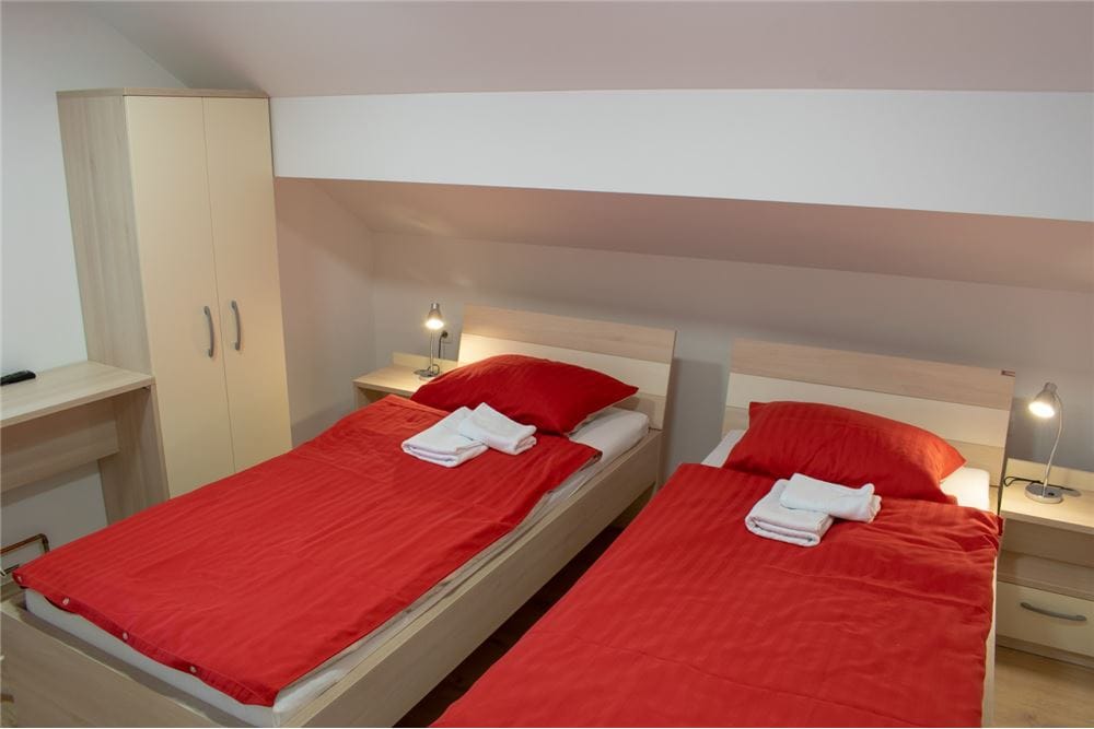 A well-lit sleeping area features two separate beds dressed in red bedding, accompanied by neatly folded white towels. Each bed has a bedside table with a lamp. A storage cabinet is positioned nearby, providing additional space for personal belongings.
