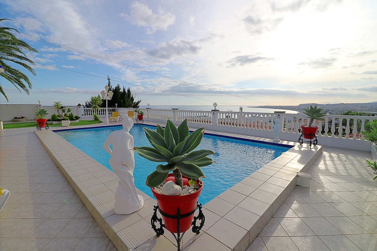 A shared swimming pool is surrounded by a tiled patio, featuring elegant planters filled with vibrant greenery. The coastline can be seen in the distance, with clouds reflecting soft light above. Palm trees are visible, contributing to a serene outdoor space.