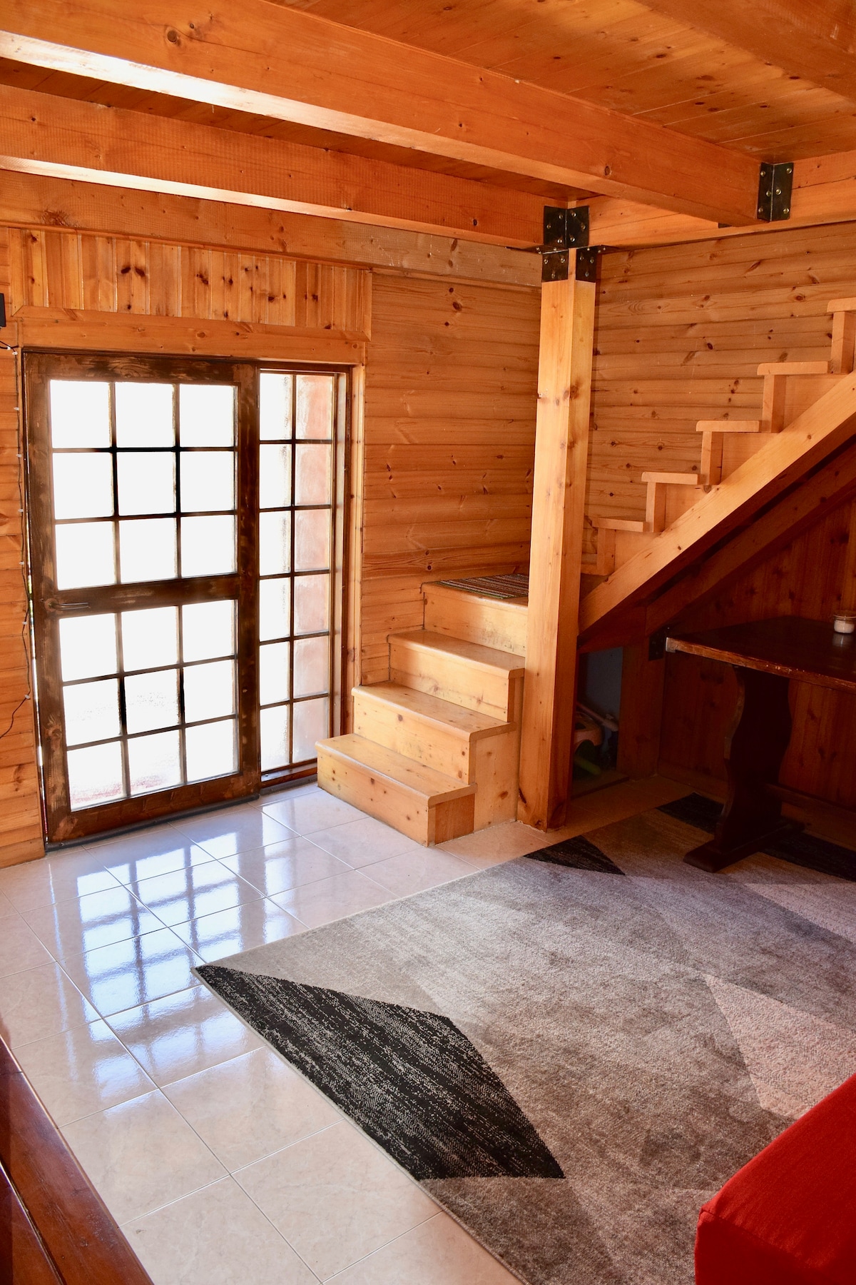 Natural wood paneling creates a warm ambiance in this interior space. A set of wooden stairs leads to the upper level, while large windows allow light to fill the room. A modern area rug adds texture to the tiled floor.