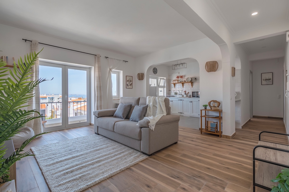 The spacious lounge area features large windows allowing natural light to illuminate the room. A comfortable sofa is positioned on a woven rug, with a cozy throw casually draped over it. A decorative plant adds a touch of greenery, complementing the modern furnishings.