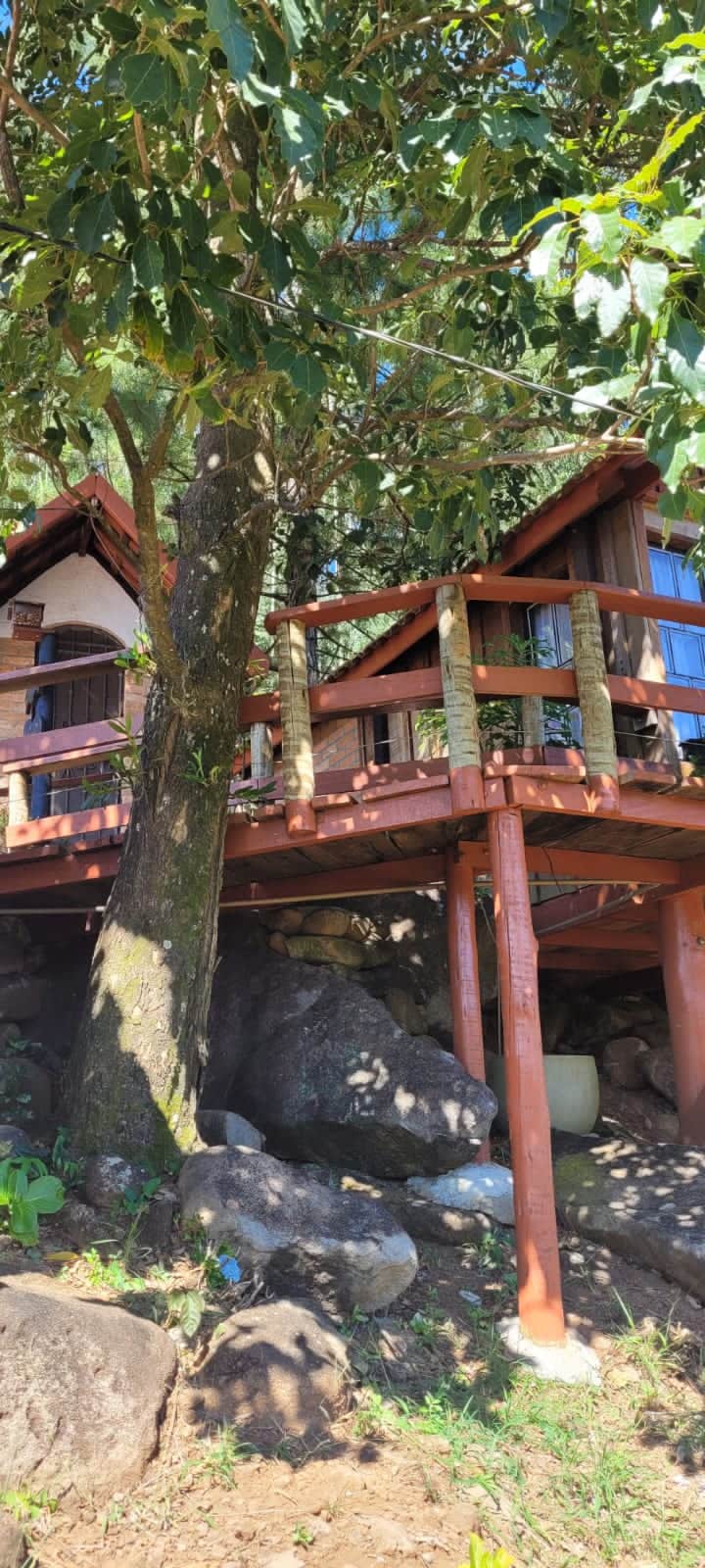 A wooden cabin perched on stilts is surrounded by lush greenery and large boulders. The structure features a sloped roof and a balcony with wooden railings, offering a natural and elevated perspective of the surroundings.