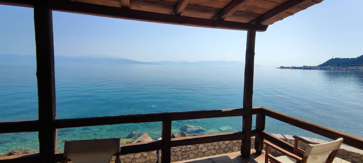A wooden balcony offers an unobstructed view of the calm, clear blue waters of the sea, with distant landmasses visible on the horizon. The serene atmosphere is enhanced by the gentle waves lapping against the rocky shoreline below.